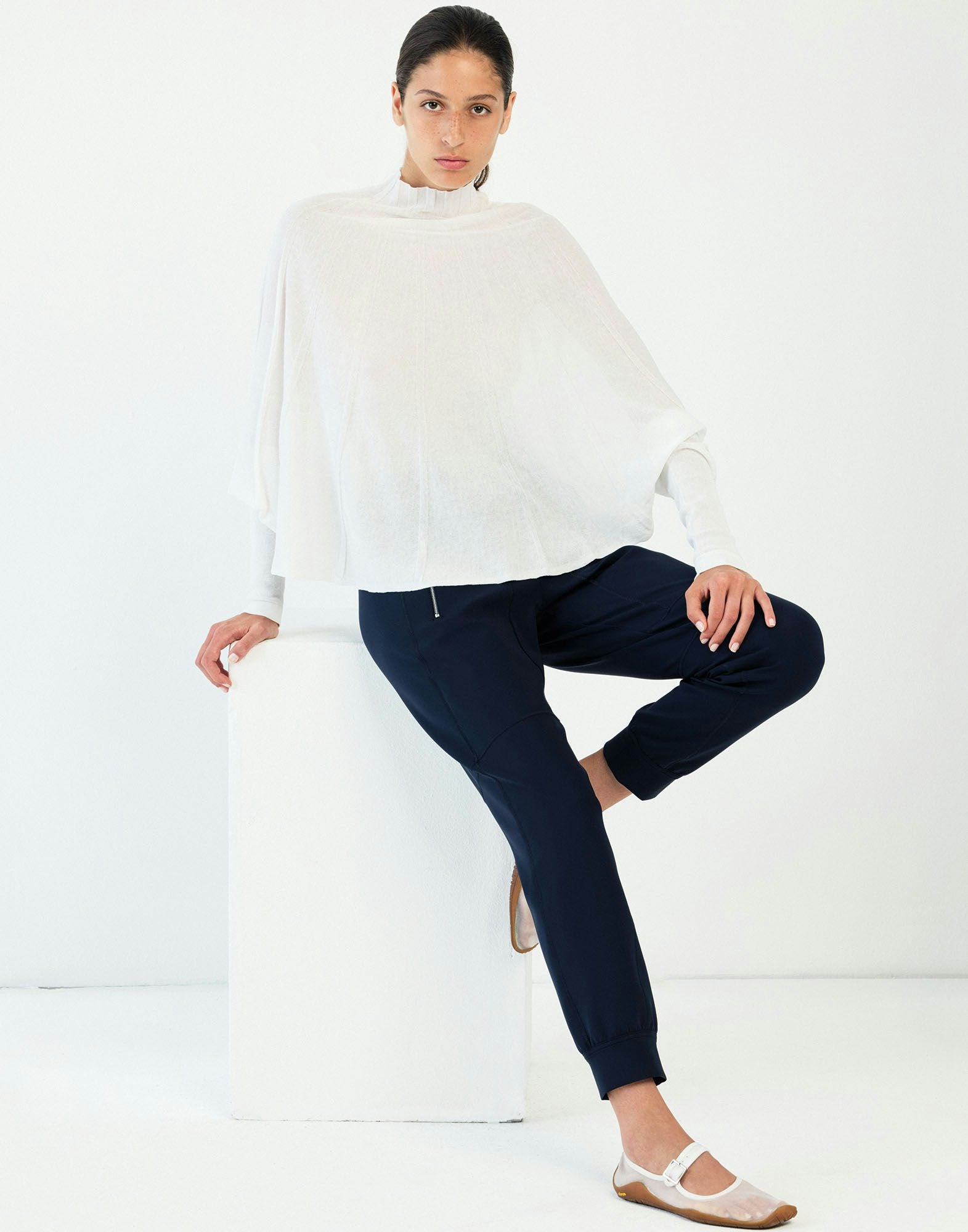 Woman sitting on a white stool wearing a white top and dark blue pants against a white background