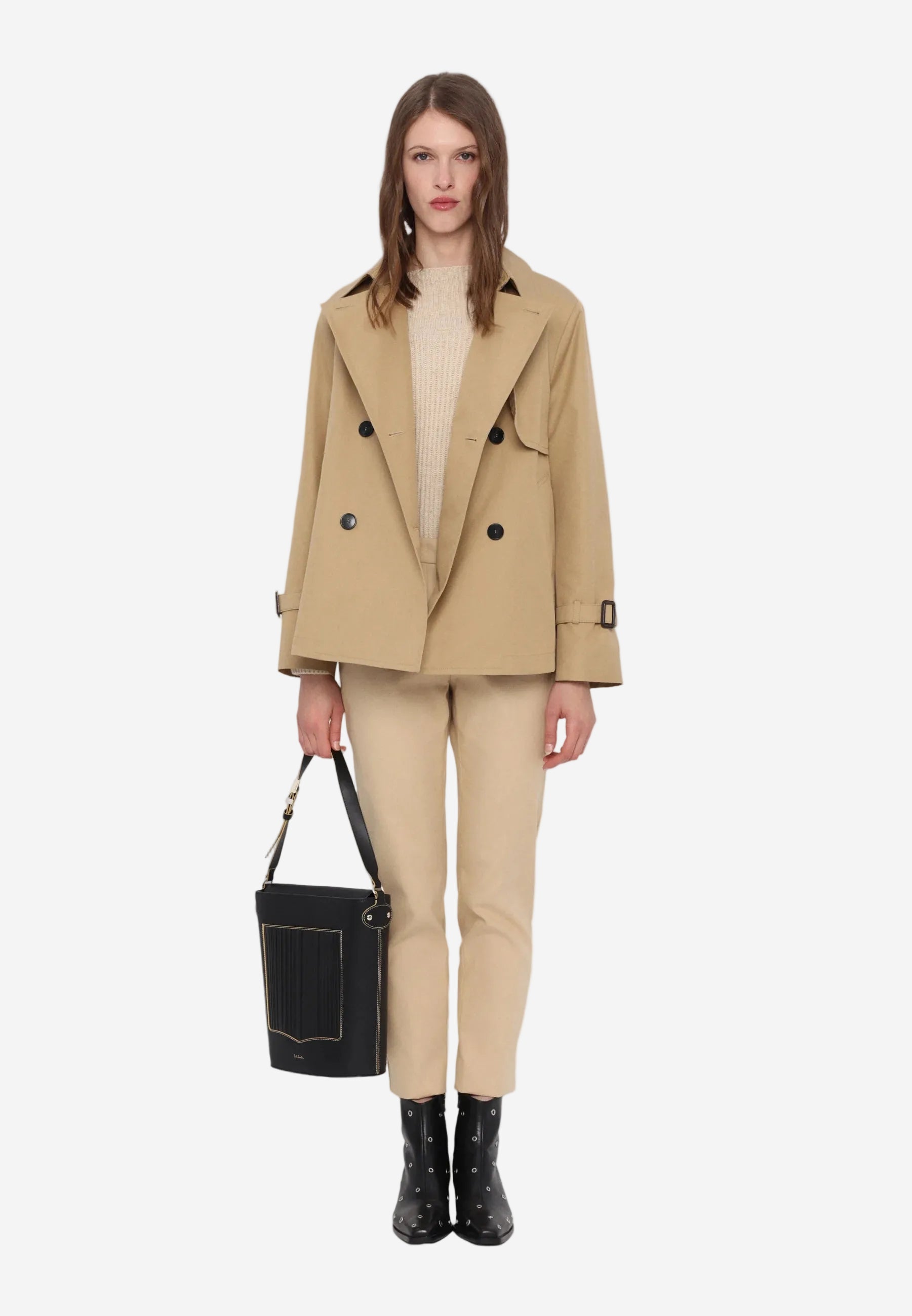 Woman wearing a beige trench coat, pants, and boots with a black handbag on a white background