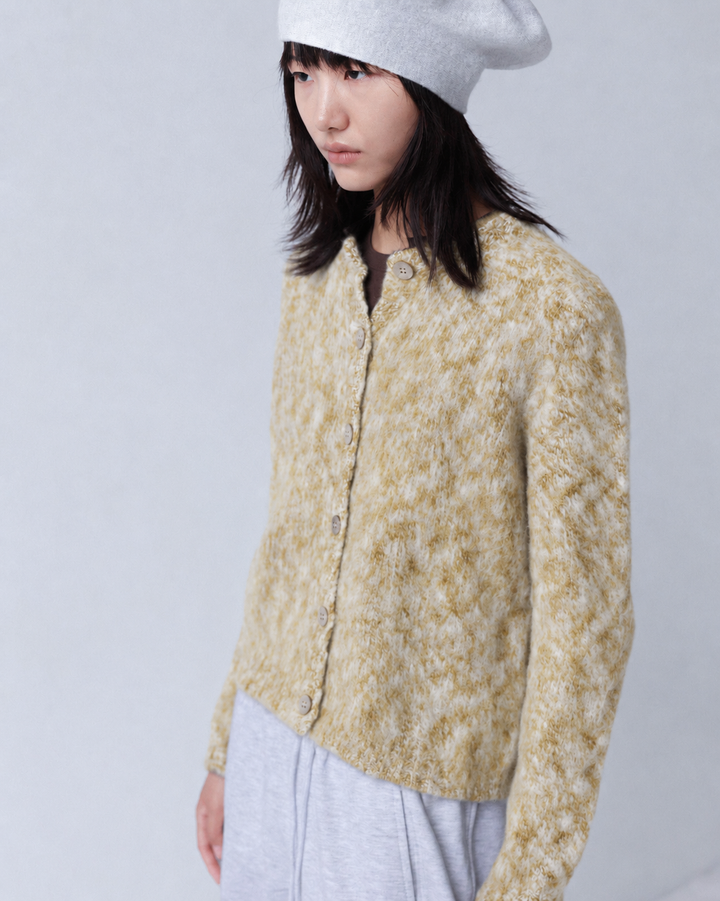A model wearing a textured, marled mustard yellow and cream knit cardigan, a light gray beanie, and light gray sweatpants, photographed against a neutral light gray studio background.