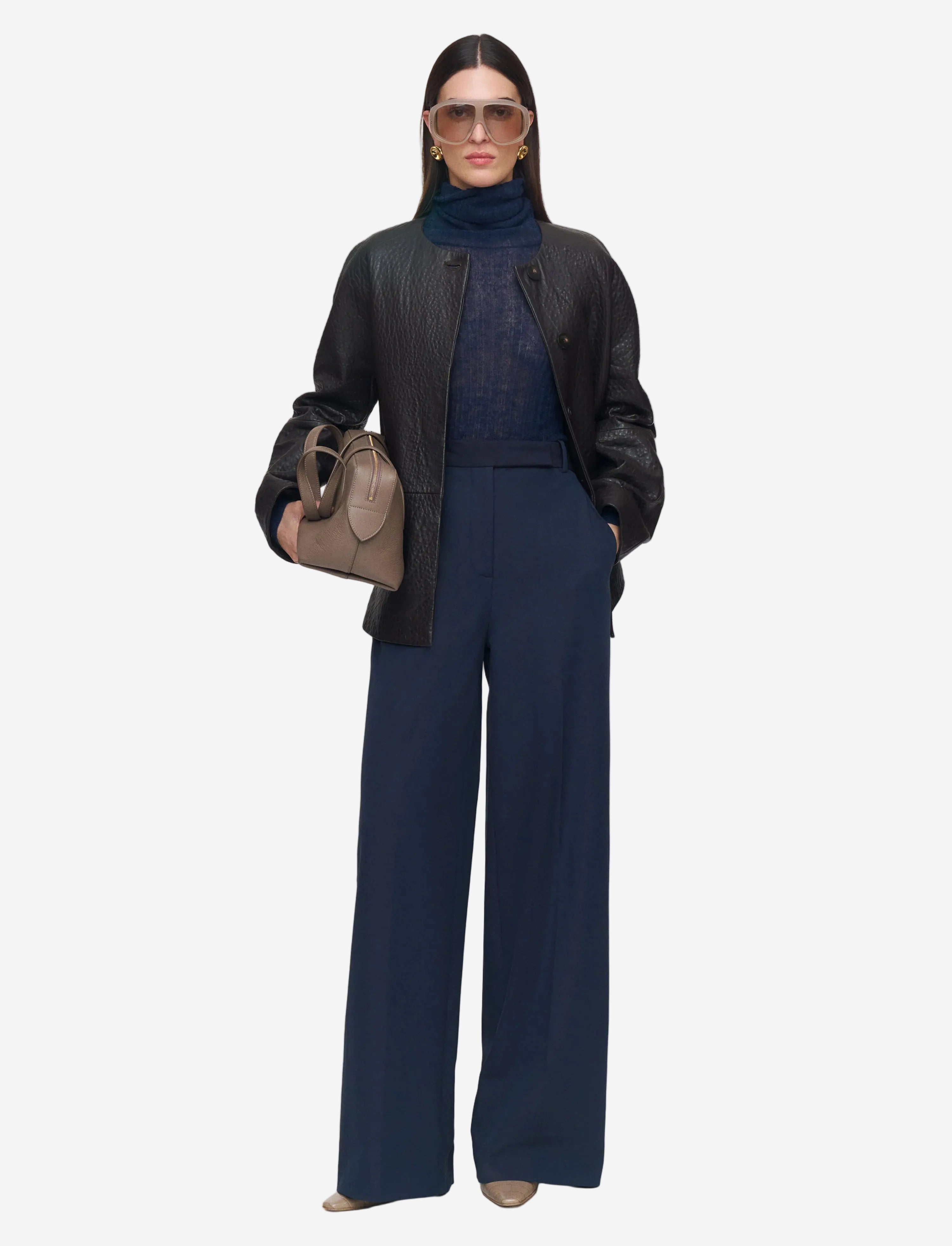Woman wearing a black leather jacket, navy turtleneck, and wide-leg pants holding a beige handbag on a white background.